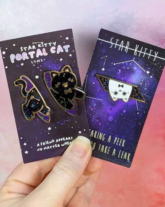A hand holds two Star Kitty Portal Cat enamel pins on their original backing cards against a pink and purple gradient background. The pins feature a black cat with a purple and blue galaxy pattern inside a triangular portal frame.