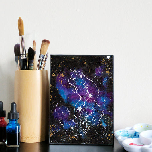 Framed artwork of a cat with a starry night sky design next to art supplies on a table.